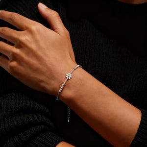 May include: A delicate silver bracelet featuring a small daisy charm with white petals and a central stone. The bracelet is adorned with small, sparkling stones along the chain. The bracelet is shown on a wrist against a dark background.