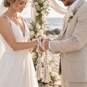 May include: A handfasting cord featuring braided beige and white fabric strands with intricate lace detailing. The cord ends in two large, textured tassels, each adorned with a small, heart-shaped wooden charm engraved with the word Love.