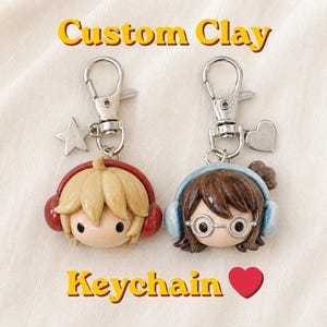 Puede incluir: Dos llaveros de arcilla personalizados con cabezas estilo chibi y cierres de mosquetón plateados. Una figura tiene cabello rubio y auriculares rojos con un dije de estrella, la otra cabello castaño, gafas y auriculares azules con un dije de corazón.