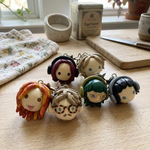 May include: A collection of five small, round, sculpted character keychains with distinct features. Each figure has a smooth, pale face with simple blue dot eyes. The characters feature varied hair colors including red, blonde, green, and black, with accessories like headphones, glasses, and piercings.
