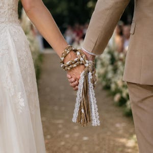 Könnte beinhalten: Ein Handfasting-Seil aus geflochtenem beigefarbenem Seil und natürlichen Jutefasern, verziert mit weißer Spitzenborte. Das Seil ist um zwei ineinandergreifende Hände gewickelt, wobei die Enden in einer quastenartigen Form mit Spitzenbändern herabhängen.