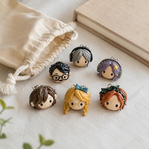 May include: Six small, sculpted character head charms with metal loops. Features include a figure with black hair and glasses, a grey-haired figure with headphones, a purple-haired figure with a star, a brown-haired figure, a blonde figure with a blue bow and glasses, and a red-haired figure with a green headband.