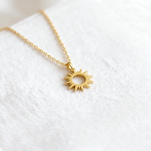 以下が含まれることがあります： 太陽のペンダントが付いたゴールドカラーのネックレス。ペンダントは、外側に伸びる尖った光線を持つ円形の中心部を備えています。チェーンとペンダントは明るく光沢のあるゴールドです。ネックレスは白くテクスチャ加工された表面に表示されています。