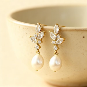 May include: A pair of gold-toned earrings with a leaf-like design. Each earring features clear, marquise-cut crystals and small round crystals, with a teardrop-shaped pearl pendant. The earrings are displayed against a neutral background.