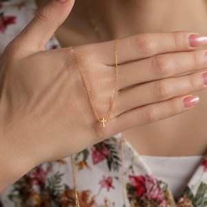 Op de afbeelding: Een delicate goudkleurige ketting met een kleine kruisvormige hanger. De ketting wordt getoond tegen een bloemrijke achtergrond. De ketting is dun en het kruis is gecentreerd, wat een eenvoudig en elegant ontwerp creëert.