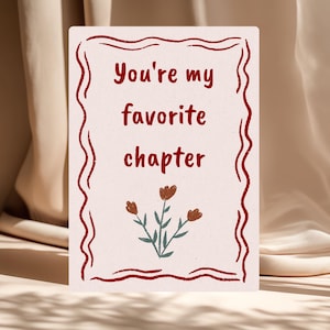 Puede incluir: Tarjeta de felicitación rectangular con el texto "You're my favorite chapter" en un estilo manuscrito. La tarjeta tiene un borde rojo ondulado y una ilustración floral en la parte inferior. El fondo es beige neutro.