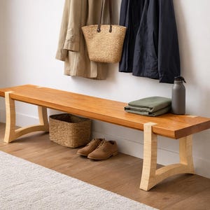 May include: A wooden bench with a natural finish, featuring light-colored legs. A woven basket, brown shoes, a green folded blanket, and a gray water bottle are on the bench. Coats and a straw tote bag hang on the wall.