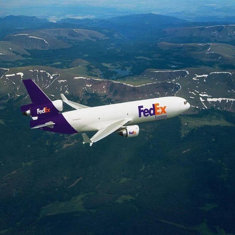 May include: A white FedEx cargo plane with purple accents flies over a mountainous landscape. The plane has the FedEx logo on the tail and fuselage. The sky is blue, and the mountains are green and brown.