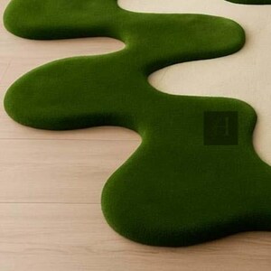 May include: A unique, organic-shaped rug in a vibrant green color. The rug has a soft, plush texture and a flowing, irregular design, resembling a stylized landscape element. The rug is placed on a light-colored wooden floor.
