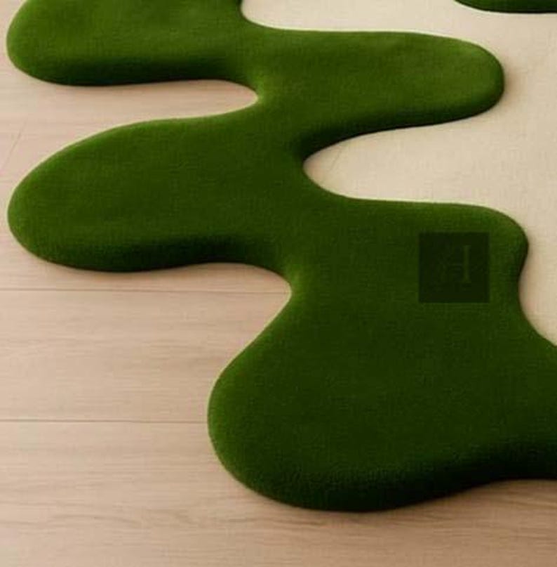 May include: A unique, organic-shaped rug in a vibrant green color. The rug has a soft, plush texture and a flowing, irregular design, resembling a stylized landscape element. The rug is placed on a light-colored wooden floor.