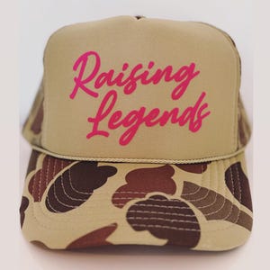 May include: A tan trucker hat with a camouflage pattern on the brim. The hat features the pink text "Raising Legends" across the front. The hat has a mesh back and a curved brim.
