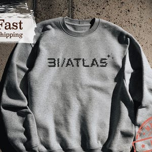 May include: Ash-colored sweatshirt with the text "3I/ATLAS" in a pixelated font. The sweatshirt has a crew neck and long sleeves. The image also includes a "Made in USA" stamp and the words "Yes Terday Club".