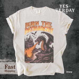 May include: Ivory-colored t-shirt featuring a dragon breathing fire, with the text "BURN THE PATRIARCHY" in orange. The graphic includes a castle, bats, and a river. The shirt has rolled-up sleeves and a crew neck.