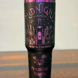 May include: Black travel mug with a black lid and a pink and purple design. The text on the mug reads "Midnight Margaritas" and "Practical Magic". The design includes a bottle, skull, and botanical illustrations.
