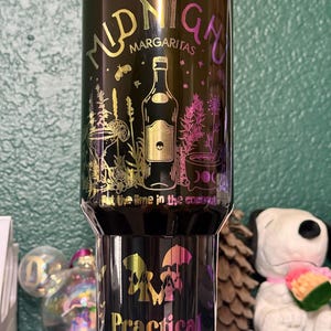 May include: A black tumbler with a metallic iridescent finish featuring the text Midnight Margaritas and Practical Magic. The design includes illustrations of a liquor bottle, a cocktail glass, a mortar and pestle, and the text Put the lime in the coconut.