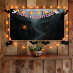 May include: A Halloween-themed display featuring a framed painting of a spooky street scene, adorned with pumpkin lights, bats, spiders, and a "Happy Haunting!" banner. A wooden bowl with greenery sits below.