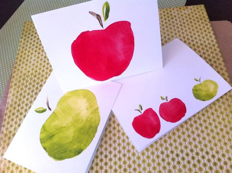May include: Three white cards with hand-painted red and green apples. The cards are blank and ready for a message.
