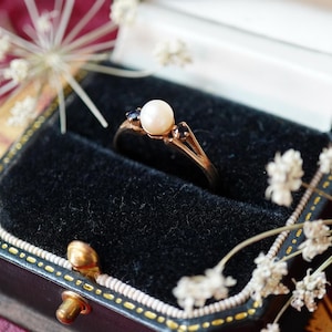 May include: A gold ring featuring a central white pearl flanked by two small dark gemstones. The ring is set within a black velvet-lined jewelry box, which is open to display the piece clearly against the dark, soft interior fabric.