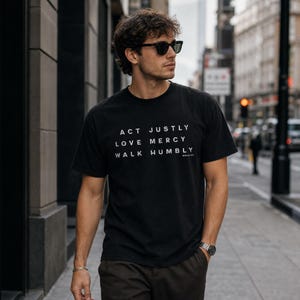 May include: A black short-sleeved t-shirt featuring white text that reads ACT JUSTLY, LOVE MERCY, WALK HUMBLY, with the smaller text MICAH 6:8 printed below. The shirt has a crew neckline and a standard fit, constructed from a solid black fabric.