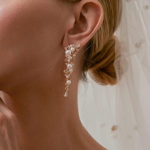 May include: A gold-coloured earring with a flowing design. The earring features clusters of pearls and clear crystals, creating a floral pattern. The earring is worn on the ear, with a blurred background.