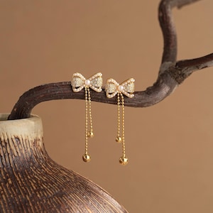 Puede incluir: Un par de pendientes dorados con forma de lazo con cadenas colgantes y detalles esféricos. Los lazos están incrustados con pequeñas piedras transparentes y una perla central. Los pendientes se exhiben en una rama marrón sobre un fondo neutro.