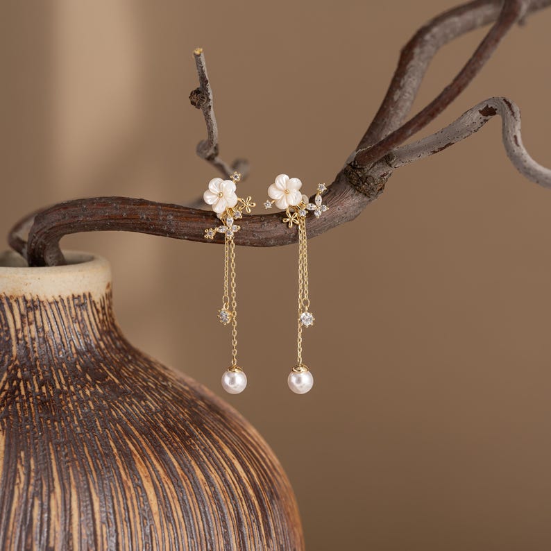 May include: A pair of gold-toned earrings featuring white flower-shaped studs with dangling chains. Each chain has a small, sparkling gem and a white pearl at the end. The earrings are displayed on a brown branch against a blurred background.