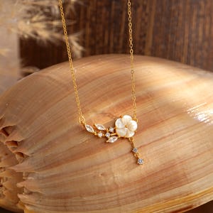 May include: A delicate gold-toned necklace featuring a white flower pendant with sparkling accents. The floral design includes leaf-shaped details and dangling crystals. The necklace is displayed on a seashell, highlighting its elegant and nature-inspired aesthetic.