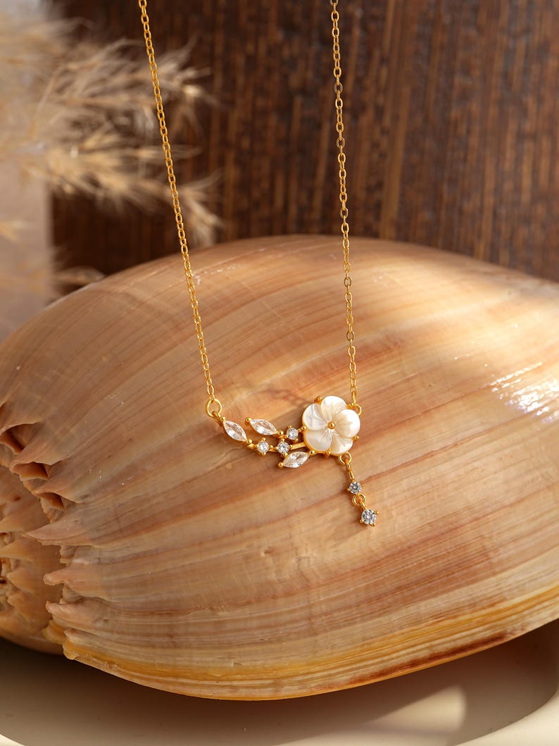 May include: A delicate gold-toned necklace featuring a white flower pendant with sparkling accents. The floral design includes leaf-shaped details and dangling crystals. The necklace is displayed on a seashell, highlighting its elegant and nature-inspired aesthetic.