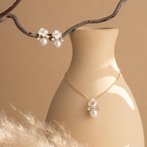 Puede incluir: Un conjunto de collar y pendientes dorados. El collar presenta un colgante con un diseño de flor blanca, una perla y pequeños cristales. Los pendientes tienen un diseño similar. Las joyas se exhiben sobre un fondo beige.
