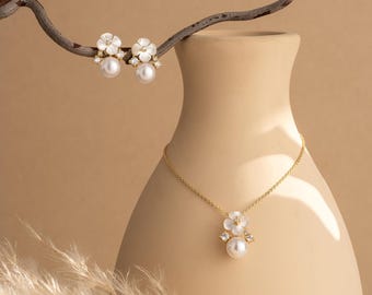 May include: A gold necklace and matching earrings set. The necklace features a pendant with a white flower design, a pearl, and small crystals. The earrings have a similar design. The jewelry is displayed on a beige background.