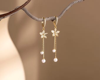 May include: A pair of gold-colored dangle earrings. Each earring features a flower-shaped charm with clear stones, a long chain, and two dangling white pearls. The earrings are suspended from a branch against a soft, neutral background.