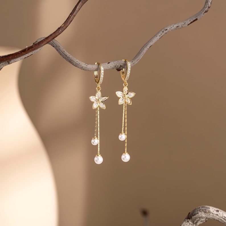 May include: A pair of gold-coloured dangle earrings. Each earring features a flower-shaped charm with clear stones, a long chain, and two dangling white pearls. The earrings are suspended from a branch against a soft, neutral background.