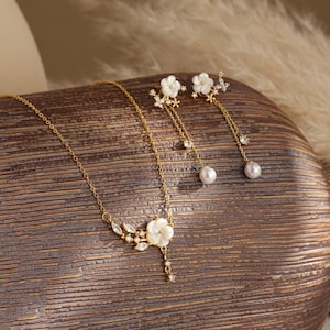 May include: A gold-toned jewelry set featuring a necklace and earrings. The necklace has a floral pendant with a pearl drop, and the earrings have a floral design with dangling pearls. The set is displayed on a wooden surface.