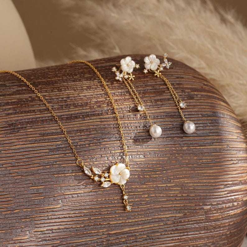 May include: A gold-toned jewelry set featuring a necklace and earrings. The necklace has a floral pendant with a pearl drop, and the earrings have a floral design with dangling pearls. The set is displayed on a wooden surface.