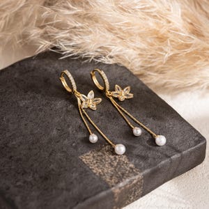 May include: Gold-coloured hoop earrings with clear crystals and dangling floral charms. Each earring has a small flower pendant and a single white pearl at the end of a delicate chain. The earrings are displayed on a dark grey surface.