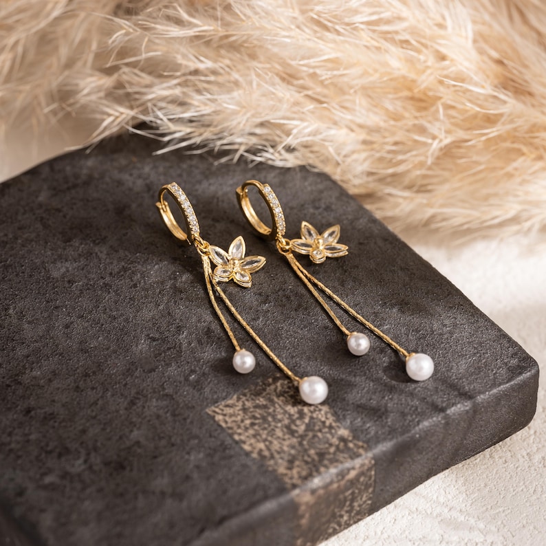 May include: Gold-coloured hoop earrings with clear crystals and dangling floral charms. Each earring has a small flower pendant and a single white pearl at the end of a delicate chain. The earrings are displayed on a dark grey surface.
