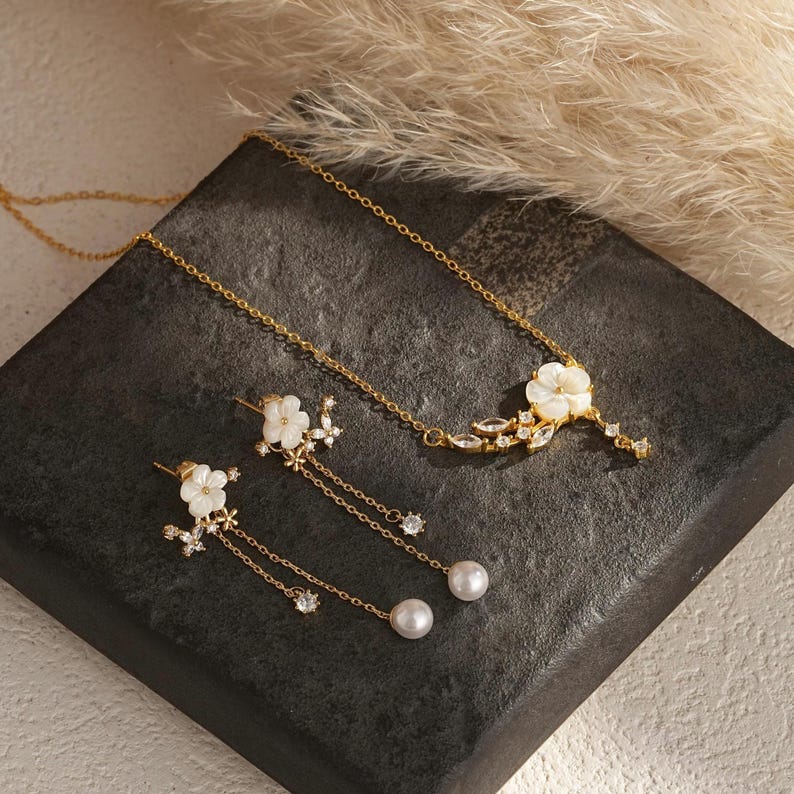 May include: A gold-toned jewelry set featuring a necklace and earrings. The necklace has a floral pendant with white petals and clear accents. The earrings have a similar floral design with dangling chains and pearl drops. The set is displayed on a dark, textured surface.