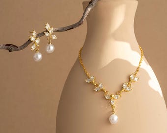 May include: A gold-toned necklace and earring set featuring leaf-shaped designs with clear crystals and dangling white pearls. The necklace has a delicate chain and a central pearl pendant. The earrings are studs with a pearl drop. The jewelry is displayed near a beige vase.