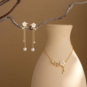 May include: Elegant gold-toned jewelry set featuring a necklace and earrings. The necklace has a floral pendant with a dangling design. The earrings are designed with a floral top, dangling chains, and pearl accents. The jewelry is displayed on a neutral background.