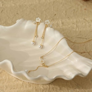 Könnte beinhalten: Ein filigranes Schmuckset mit einer goldenen Halskette und Ohrringen. Die Halskette und die Ohrringe haben ein Blumendesign mit weißen Blütenblättern und kleinen, klaren Kristallen. Die Ohrringe hängen mit einer Perle am Ende. Das Schmuckstück wird auf einer weißen, muschelförmigen Schale präsentiert.