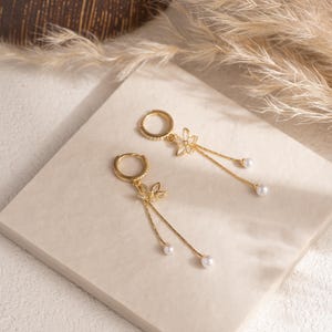 May include: A pair of gold-coloured hoop earrings featuring a floral design and dangling faux pearls. The earrings are displayed on a neutral surface, with dried ornamental grass in the background.