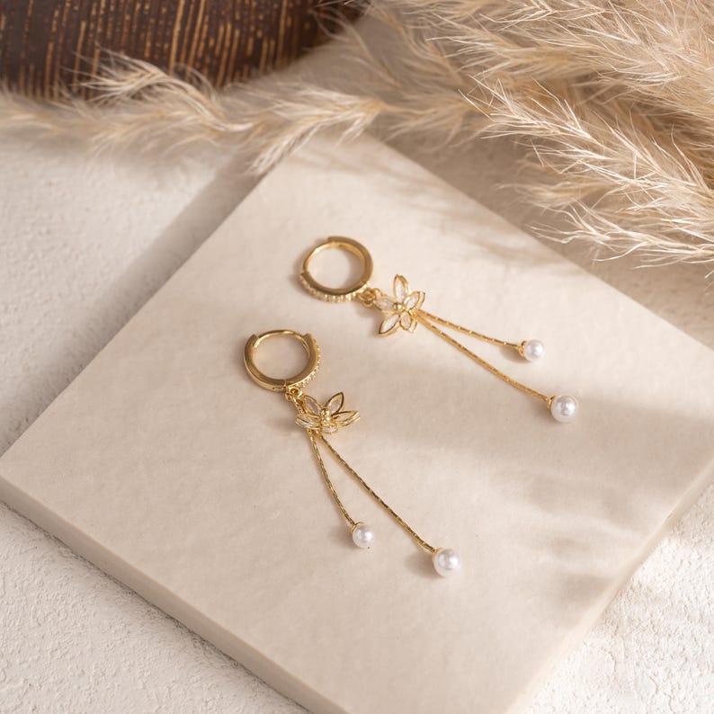 May include: A pair of gold-coloured hoop earrings featuring a floral design and dangling faux pearls. The earrings are displayed on a neutral surface, with dried ornamental grass in the background.