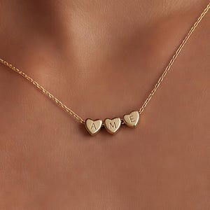 Può includere: Una collana color oro con una catenina sottile e tre piccoli ciondoli a forma di cuore. Ogni ciondolo a cuore è inciso con una lettera maiuscola, formando la sequenza A, M ed E da sinistra a destra al centro della catenina.