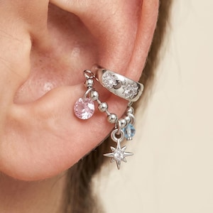 May include: Silver ear cuff featuring a curved band with embedded clear stones and a dangling chain. The chain holds a pink gemstone, a blue gemstone, and a star-shaped charm. The cuff is worn on the ear.