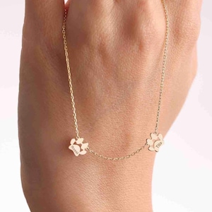 May include: A gold-coloured chain necklace featuring two small, flat paw print charms. Each charm is engraved with a name, with one reading 'Dobby' and the other reading 'Tito'. The delicate metal chain connects the two charms in a symmetrical design.