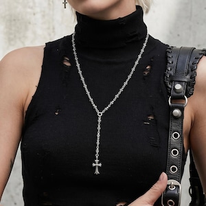 Puede incluir: Un collar con colgante de cruz plateada con un diseño en forma de Y, usado con pendientes de cruz a juego. La modelo lleva un top negro sin mangas de cuello alto y una pulsera plateada. El look es atrevido y elegante.