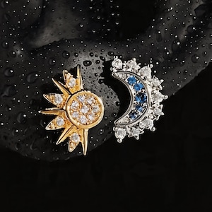 Pode incluir: Um par de brincos com tema celestial. Um brinco é um design de sol dourado com pedras transparentes. O outro é uma lua crescente prateada com pedras azuis e transparentes. Os brincos estão em um fundo escuro.