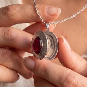 May include: A silver-toned oval locket pendant on a delicate chain. The locket is open, revealing a dark red gemstone set into the outer cover and a small black and white portrait photograph of a person visible inside the metal frame.