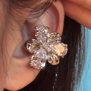 Puede incluir: Un pendiente tipo ear cuff de color plateado con forma de flor de cinco pétalos. Cada pétalo presenta una gran piedra de circonita cúbica transparente en forma de pera. El centro de la flor está acentuado con un grupo de circonitas redondas y claras.