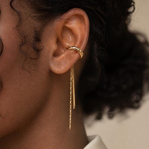 May include: A gold ear cuff and chain earring set. The ear cuff curves around the upper ear, while the earring features a delicate chain and a straight bar that dangles down. The jewelry is shown on a person's ear.